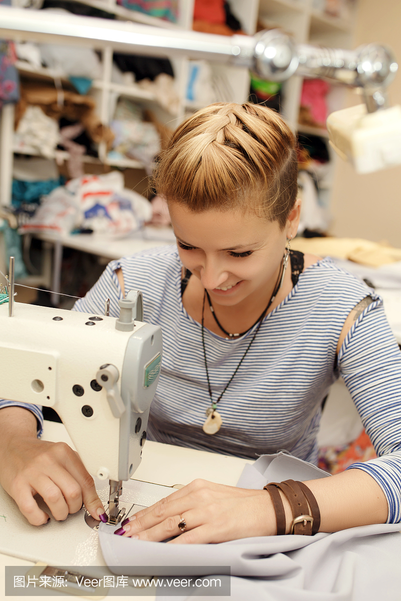 年轻的金发女裁缝布设计师在缝纫机上工作。在她的工作空间。服装制造业
